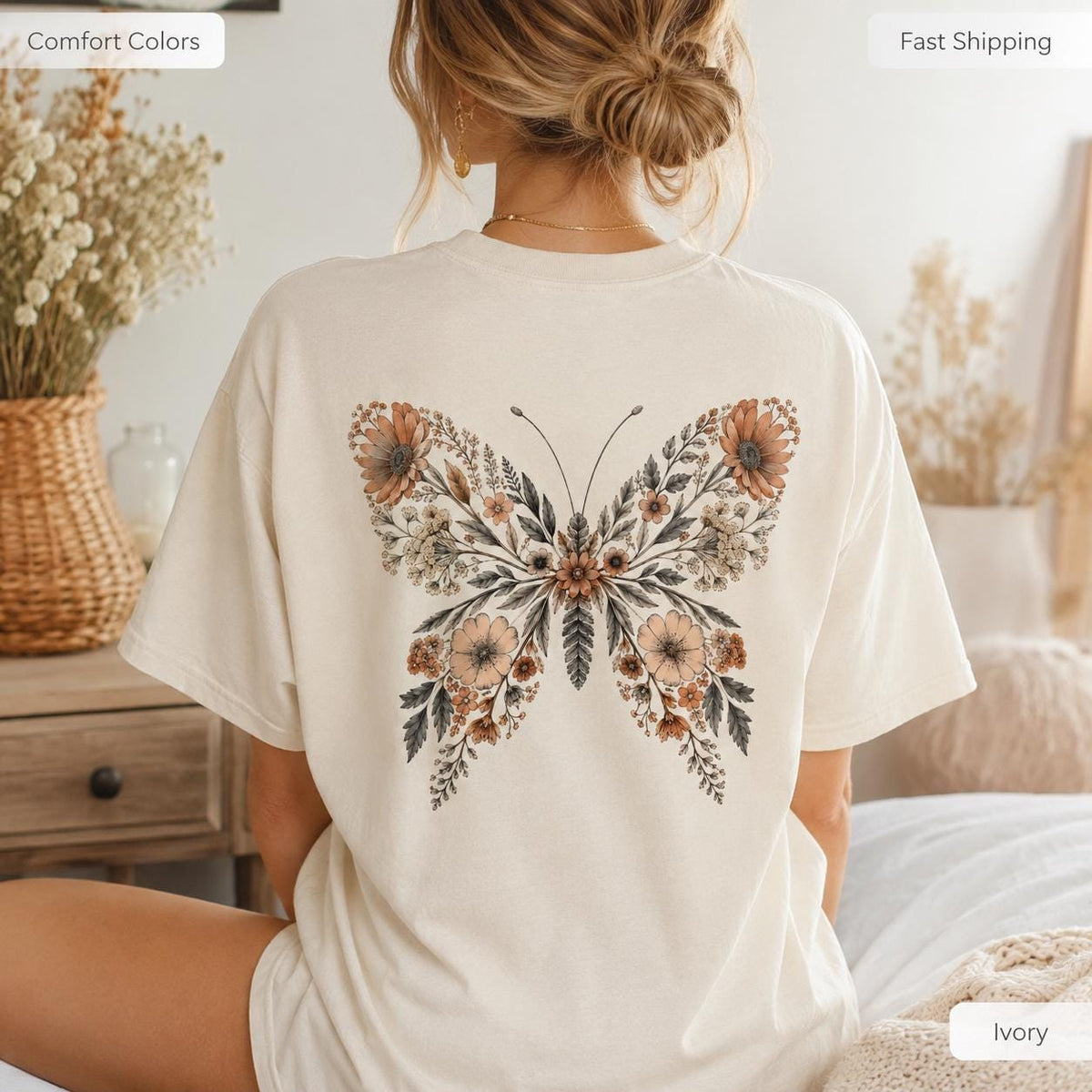 May include: An ivory short-sleeve t-shirt featuring a large butterfly graphic on the back. The butterfly wings are composed of intricate floral and botanical illustrations in shades of muted orange, brown, and dark gray, creating a detailed pattern.