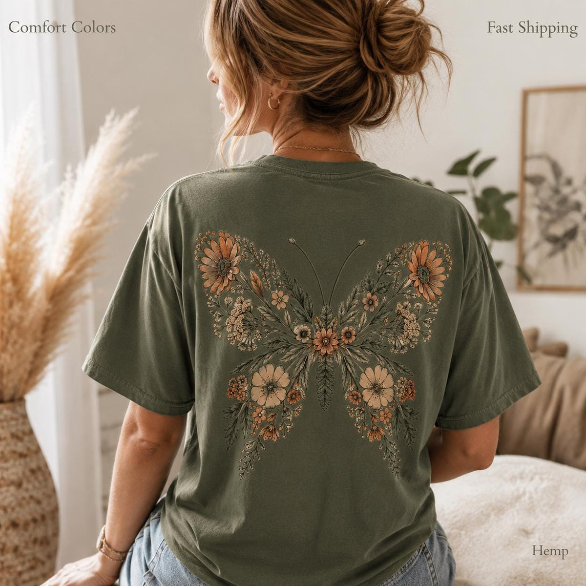 May include: An olive green short-sleeve t-shirt featuring a large graphic on the back. The graphic depicts a butterfly composed of various orange, brown, and cream-colored flowers, leaves, and stems arranged to form the shape of butterfly wings.
