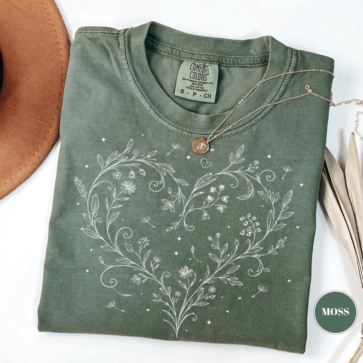 May include: A moss green t-shirt with a white floral heart design. The shirt has a crew neck and short sleeves. A gold necklace with a pendant rests on the shirt. The tag reads "Comfort Colors".