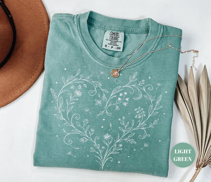 May include: Light green t-shirt with a white floral heart design. The shirt has a crew neck and short sleeves. A silver necklace with a small pendant rests on the shirt. The shirt is folded and placed on a white surface.