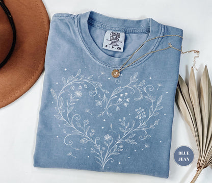 May include: A blue jean colored t-shirt with a white floral heart design. The shirt has a crew neck and short sleeves. A gold necklace with a pendant is draped over the shirt. The tag reads "Comfort Colors".