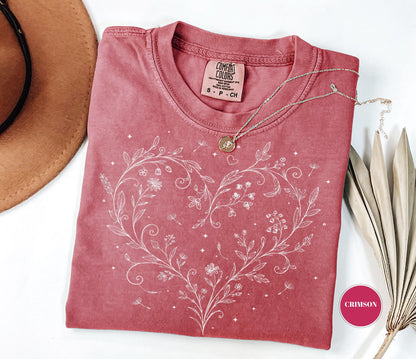 May include: Crimson-colored t-shirt with a white floral heart design. The shirt has a crew neck and short sleeves. A gold necklace with a pendant rests on the shirt. The shirt is labeled "Comfort Colors" and is size S-P-CH.