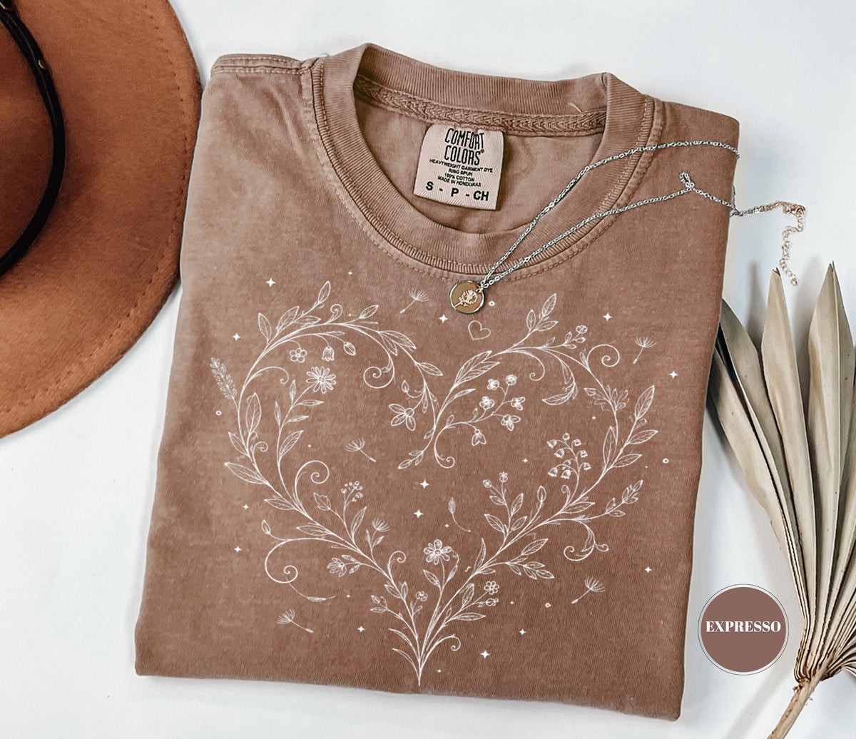 May include: A brown t-shirt with a white floral heart design. The shirt has a crew neck and short sleeves. A silver necklace with a small pendant rests on the shirt. The shirt is labeled "Comfort Colors".