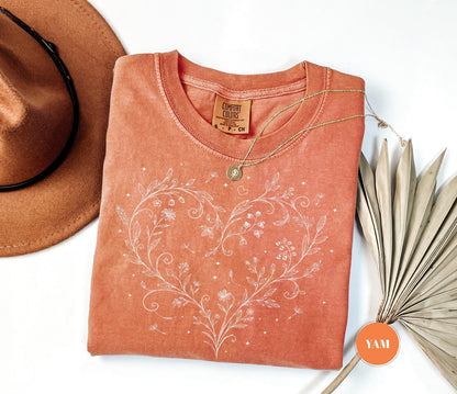 May include: Coral-colored Comfort Colors t-shirt with a white floral heart design. A gold necklace with a pendant rests on the shirt. A brown hat and dried palm leaves are also in the image. The shirt has a round neck and short sleeves.
