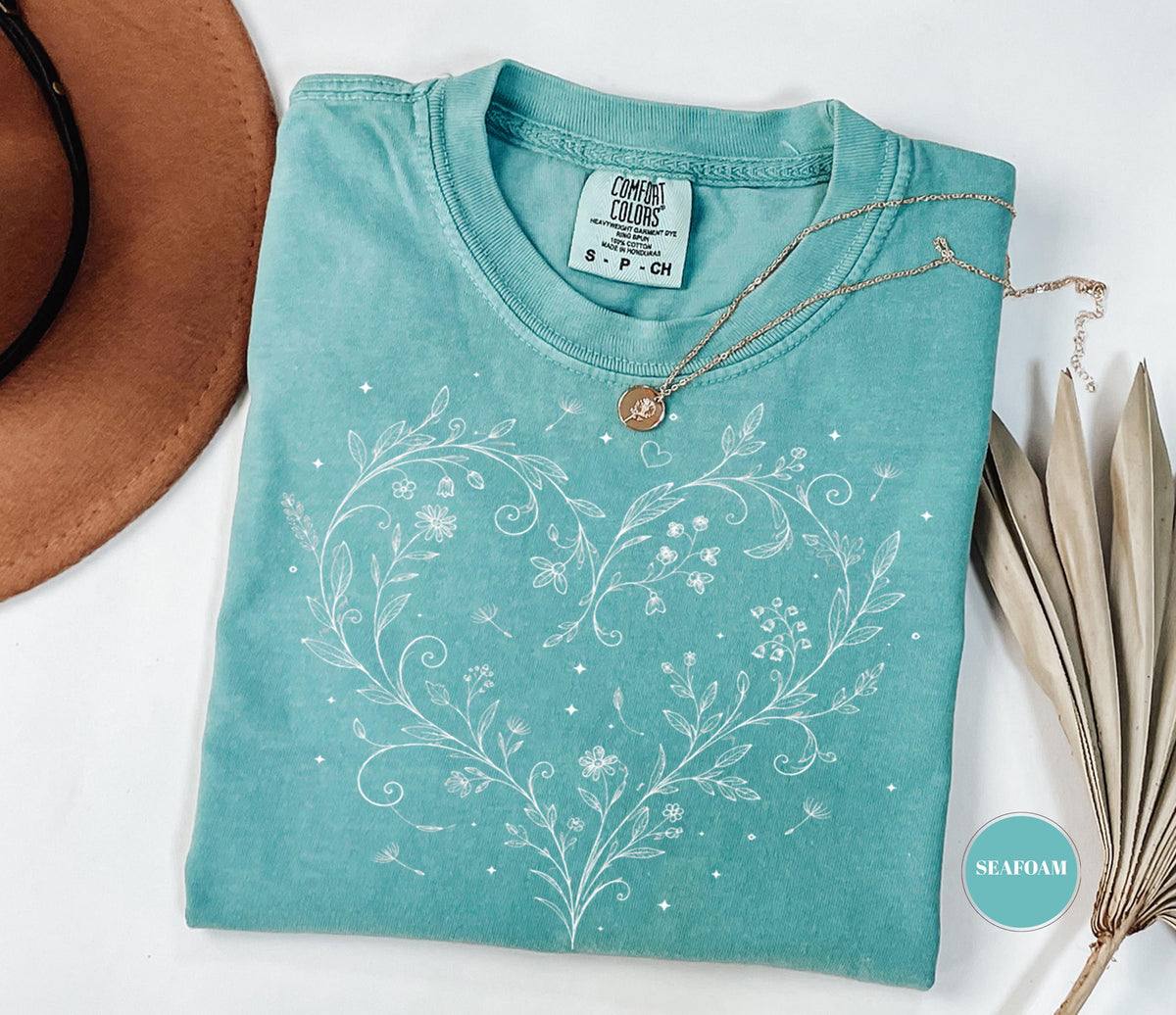 May include: Seafoam green t-shirt with a white floral heart design. The shirt has a crew neck and short sleeves. A gold necklace with a pendant rests on the shirt. The shirt is labeled "Comfort Colors".