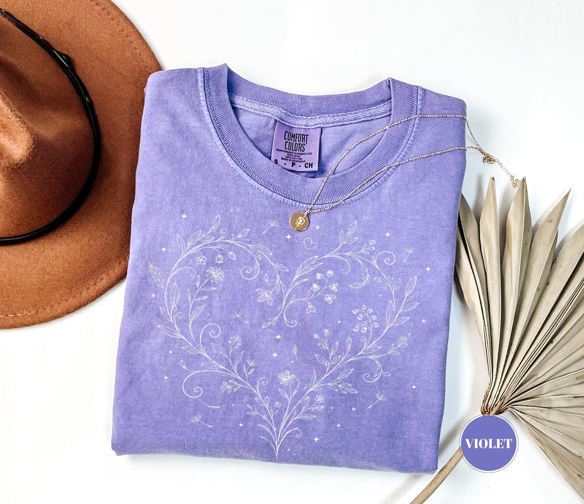 May include: A violet-colored t-shirt with a white floral heart design. The shirt has a crew neck and short sleeves. A gold necklace with a pendant is draped on the shirt. A brown hat is in the upper left corner.