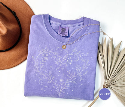 May include: A violet-colored t-shirt with a white floral heart design. The shirt has a crew neck and short sleeves. A gold necklace with a pendant is draped on the shirt. A brown hat is in the upper left corner.