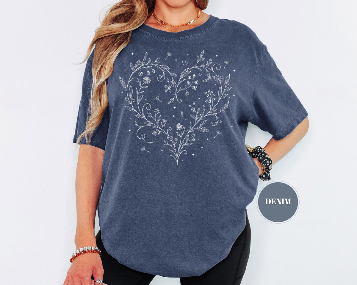 May include: A denim blue t-shirt featuring a white floral heart design. The design includes flowers, leaves, and small stars. The shirt has short sleeves and a relaxed fit. The word "DENIM" is in a gray circle.