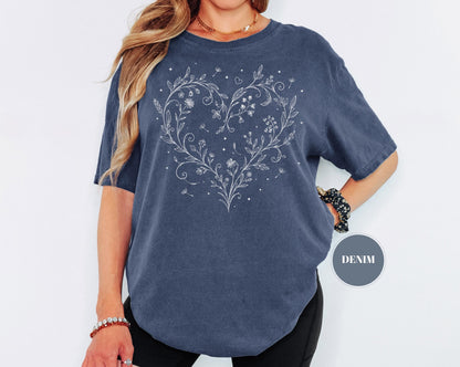 May include: A denim blue t-shirt featuring a white floral heart design. The design includes flowers, leaves, and small stars. The shirt has short sleeves and a relaxed fit. The word "DENIM" is in a gray circle.
