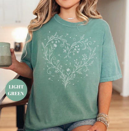 May include: Light green t-shirt with a white floral heart design. The heart is composed of delicate flowers, leaves, and small details. The shirt has short sleeves and a relaxed fit. The text "LIGHT GREEN" is in a circle on the bottom left.