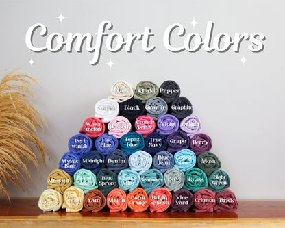 May include: A pyramid of rolled-up t-shirts in various colors, including white, black, and blue. The text "Comfort Colors" is displayed above the shirts. The shirts are arranged on a wooden surface.