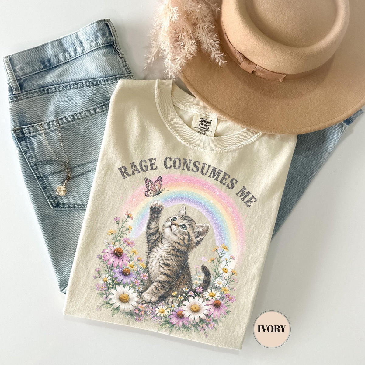 May include: Ivory-colored t-shirt with the text "RAGE CONSUMES ME" and a graphic of a kitten reaching for a butterfly in front of a rainbow. The shirt is paired with light blue jeans, a necklace, and a tan hat.