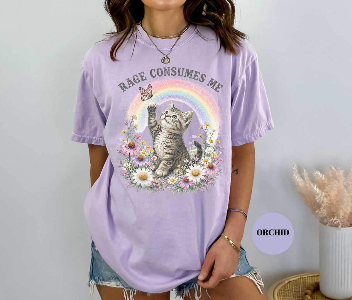 May include: A light purple t-shirt with the text "RAGE CONSUMES ME" above a graphic of a kitten reaching for a butterfly near a rainbow. The kitten is surrounded by flowers.