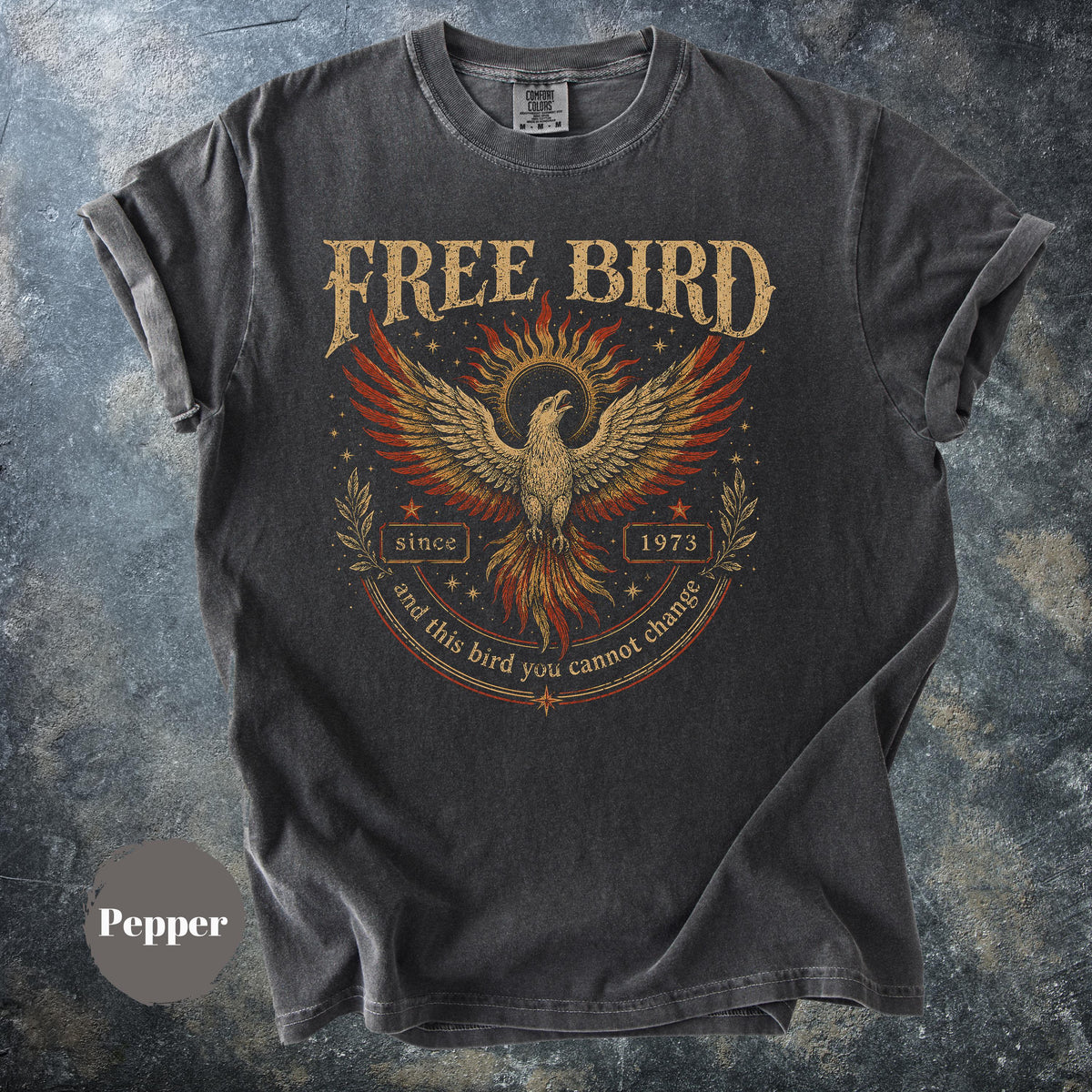 May include: Pepper-colored t-shirt with a distressed graphic of a phoenix with outstretched wings, surrounded by text that reads "FREE BIRD" above and "since 1973 and this bird you cannot change" below. The shirt has rolled-up sleeves.