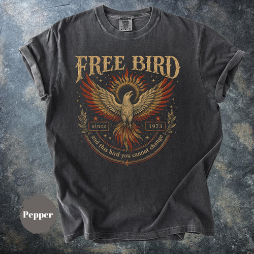 May include: Pepper-colored t-shirt with a distressed graphic of a phoenix with outstretched wings, surrounded by text that reads "FREE BIRD" above and "since 1973 and this bird you cannot change" below. The shirt has rolled-up sleeves.