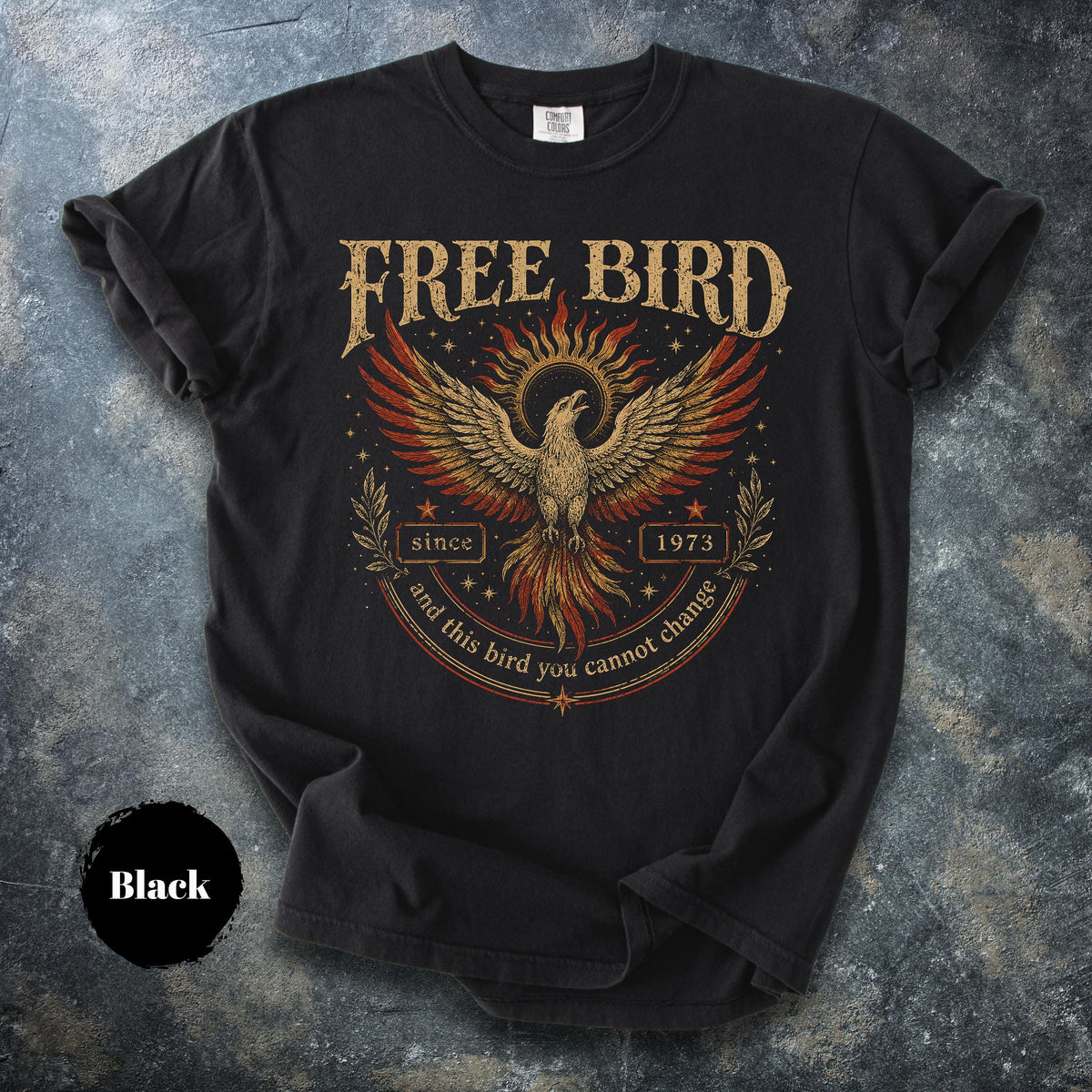 May include: Black t-shirt with a distressed graphic of a phoenix with outstretched wings, the text "FREE BIRD" above, and "since 1973" below. The design features red, gold, and brown colors. The shirt is a crew neck.