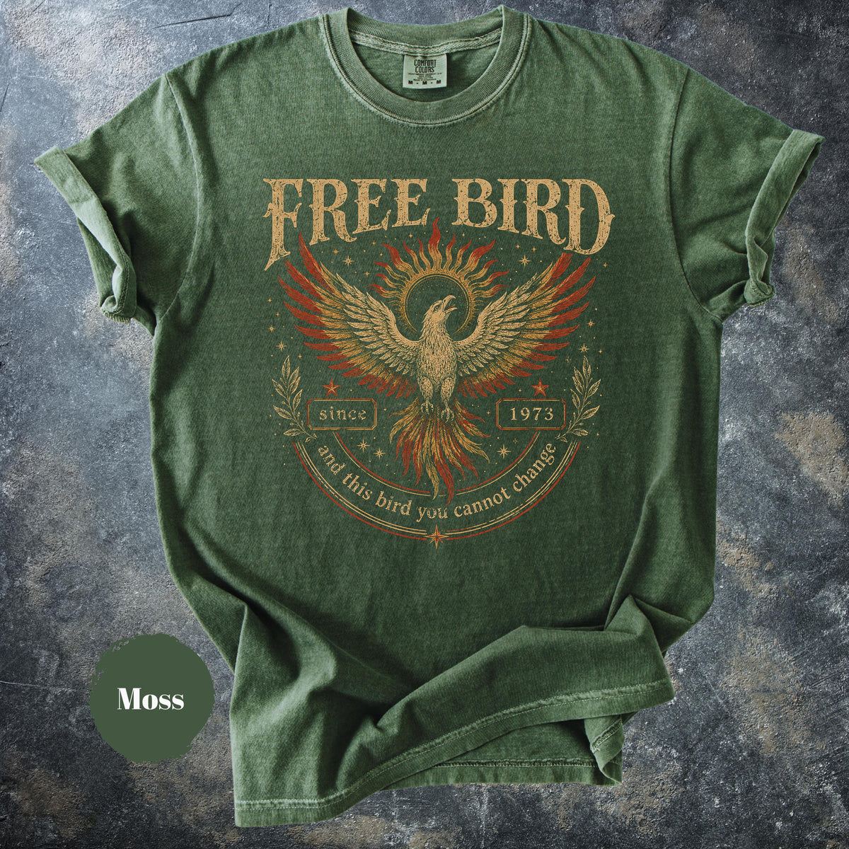 May include: Moss green t-shirt with a distressed graphic featuring a phoenix with outstretched wings, the words "FREE BIRD" and "since 1973". The text "and this bird you cannot change" is arched below the phoenix. The shirt has rolled-up sleeves.