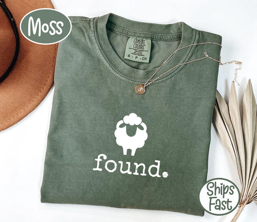May include: A moss green t-shirt with a white sheep graphic and the word "found" in white. The shirt has a crew neck and short sleeves. A gold necklace with a small pendant is draped on the shirt.