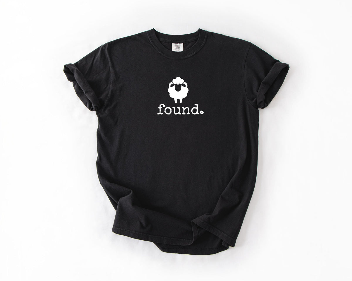 May include: Black t-shirt with a white sheep graphic and the word "found." printed below. The sheep graphic is a simple outline with a fluffy head. The shirt is a basic crew neck.