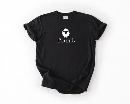 May include: Black t-shirt with a white sheep graphic and the word "found." printed below. The sheep graphic is a simple outline with a fluffy head. The shirt is a basic crew neck.