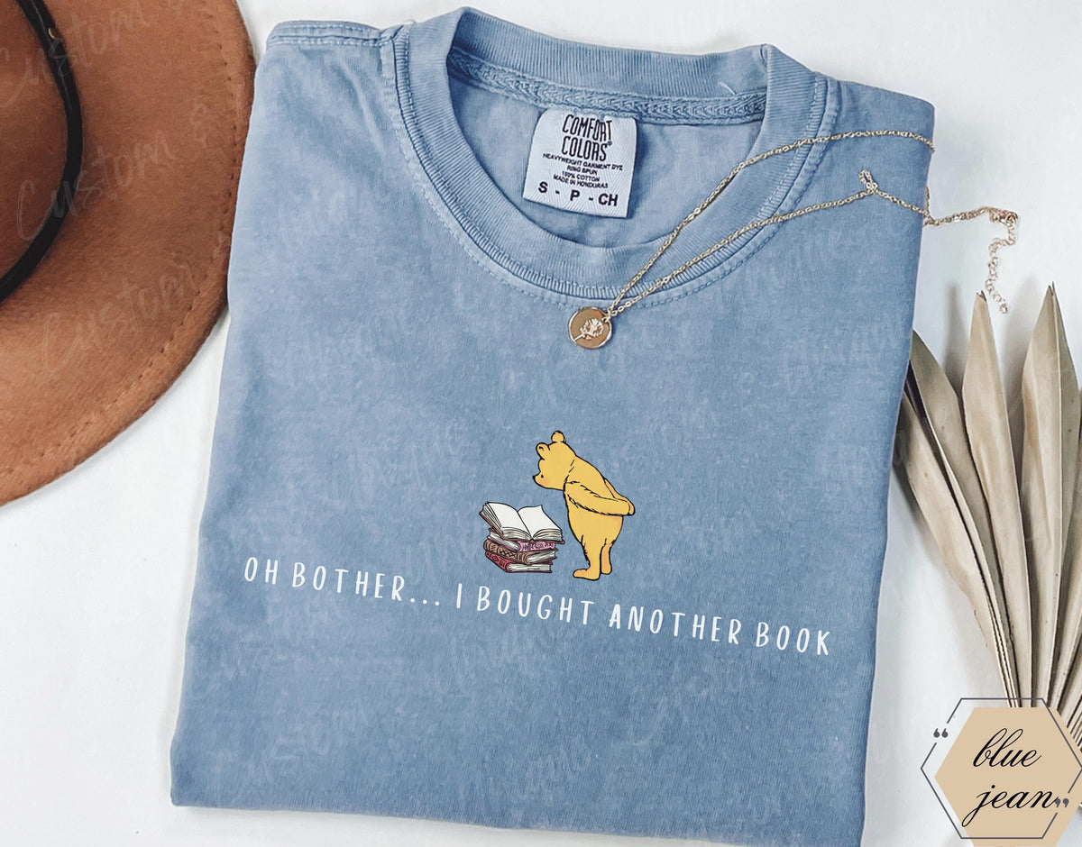 May include: A light blue Comfort Colors t-shirt featuring a graphic of Winnie the Pooh and books, with the text "OH BOTHER... I BOUGHT ANOTHER BOOK." A gold necklace rests on the shirt. The shirt is a size small.