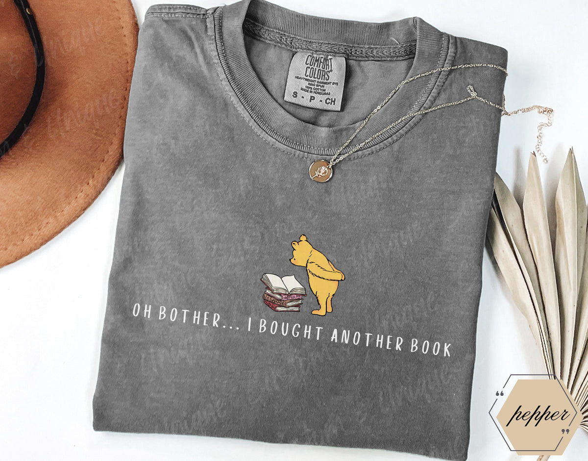 May include: A gray Comfort Colors t-shirt featuring a graphic of Winnie the Pooh with books and the text "OH BOTHER... I BOUGHT ANOTHER BOOK." The shirt has a crew neck and short sleeves. A gold necklace is draped across the shirt.