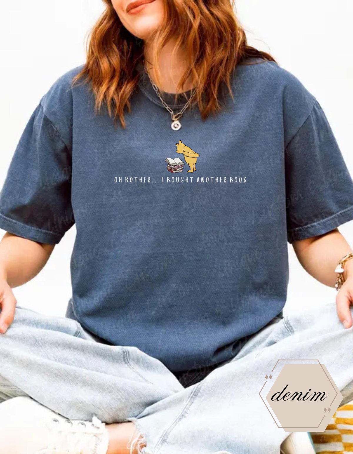 May include: A denim blue t-shirt featuring a graphic of a yellow bear and a stack of books. The text "OH BOTHER... I BOUGHT ANOTHER BOOK" is printed below the graphic. The shirt is a relaxed fit.