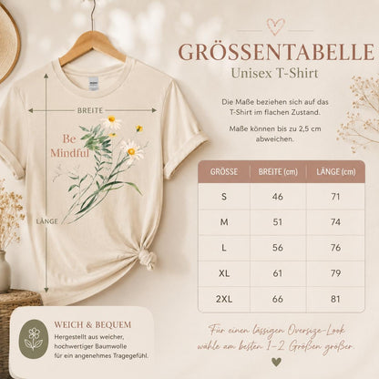 May include: A beige short-sleeved t-shirt featuring a graphic of white daisies and green foliage with the text Be Mindful. A size chart displays measurements in centimeters for sizes S through 2XL, indicating widths from 46 to 66 cm and lengths from 71 to 81 cm.