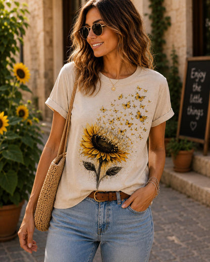 May include: A beige short-sleeve t-shirt featuring a large graphic of a yellow sunflower with a cluster of small yellow butterflies appearing to fly upward from the flower petals. The shirt has a crew neckline and rolled sleeve cuffs.