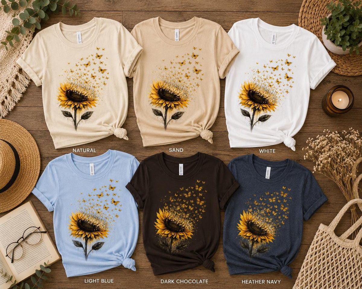 May include: Six short-sleeve t-shirts in natural, sand, white, light blue, dark chocolate, and heather navy colors. Each shirt features a graphic of a yellow sunflower with small orange butterflies flying upward from the petals in a swarm.