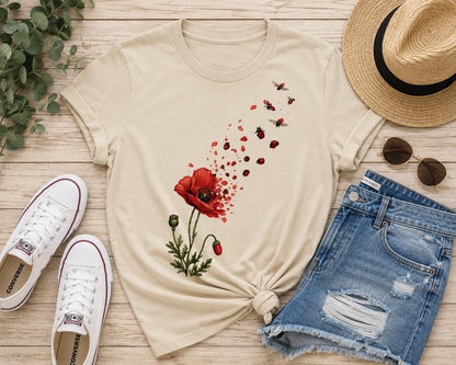 May include: A beige short-sleeve t-shirt featuring a graphic of a red poppy flower with petals transforming into a swarm of small ladybugs. The design is centered on the chest, showcasing green stems and buds at the base of the vibrant red flower.