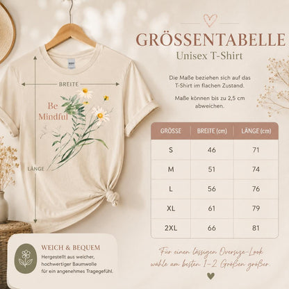 May include: A beige short-sleeved t-shirt featuring a graphic of white daisies and green foliage with the text Be Mindful. A size chart displays measurements in centimeters for sizes S through 2XL, indicating widths from 46 to 66 cm and lengths from 71 to 81 cm.