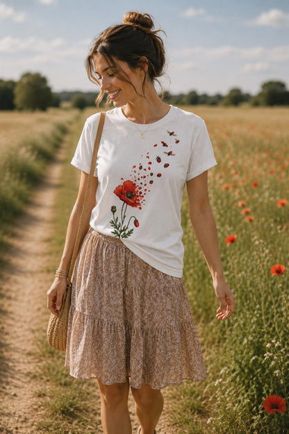 May include: A white short-sleeved t-shirt featuring a graphic of a red poppy flower with petals transforming into small ladybugs that fly upward. The shirt is paired with a tan tiered skirt with a small white floral pattern and a woven shoulder bag.