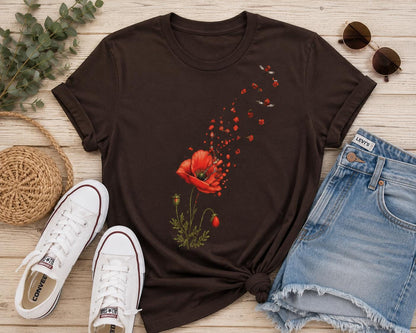 May include: A dark brown short-sleeve t-shirt featuring a graphic print of a red poppy flower with petals appearing to drift upward and away. The shirt is displayed alongside white canvas sneakers, blue denim shorts, and brown sunglasses.