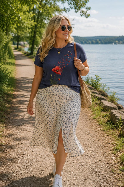 May include: A navy blue short-sleeve t-shirt featuring a red poppy flower graphic with small flying insects, paired with a cream-colored midi skirt with a blue floral print and a front slit, worn with white sneakers and a woven straw shoulder bag.