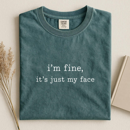 May include: A dark teal crew neck t-shirt with the white text "i'm fine, it's just my face" printed on the center chest. The shirt features a ribbed collar and a Comfort Colors brand label on the inside neck area, showcasing a solid color design.