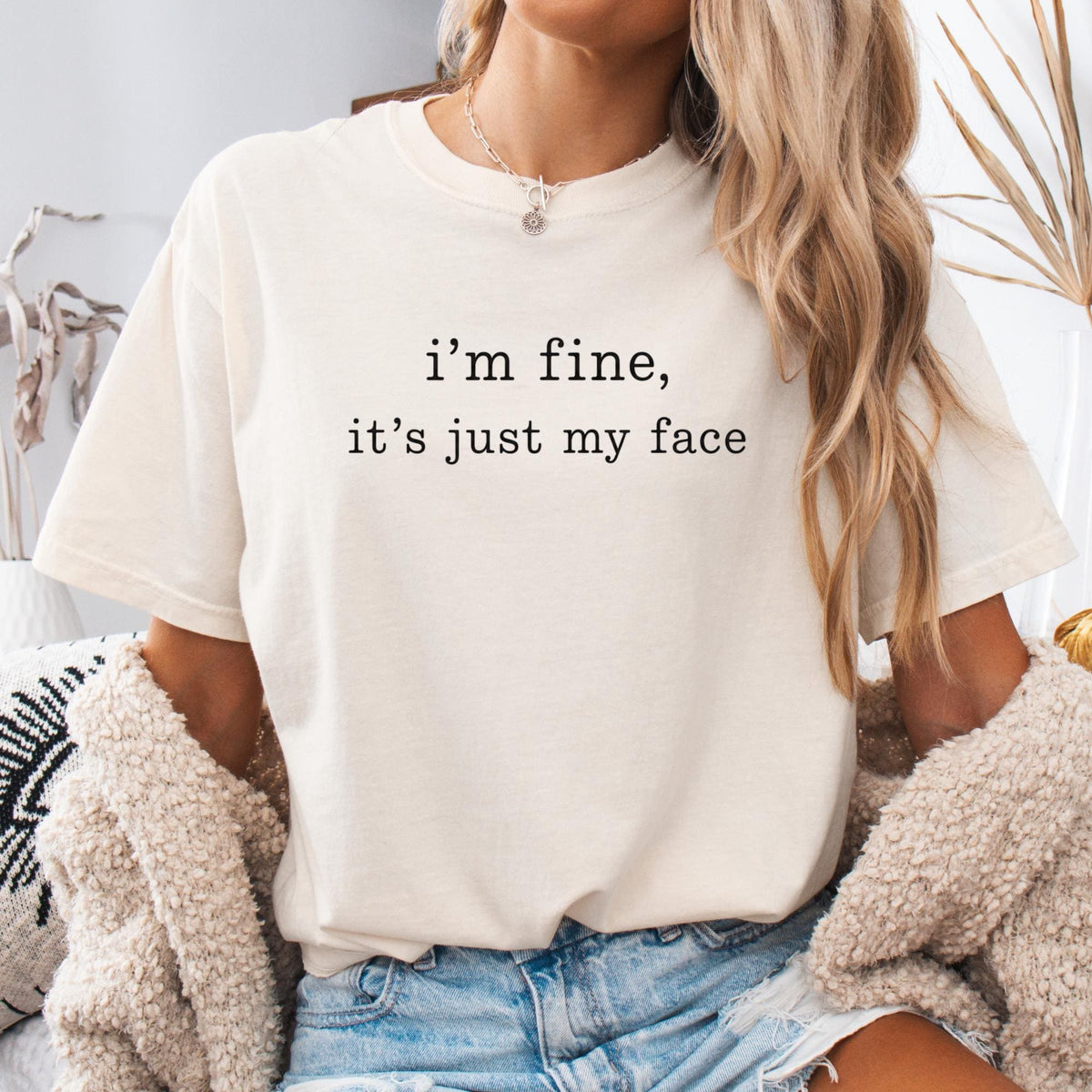 May include: A cream-colored short-sleeve t-shirt featuring the black printed text "i'm fine, it's just my face" centered on the chest. The shirt has a crew neckline and a relaxed fit, paired with light blue denim jeans and a textured beige cardigan.