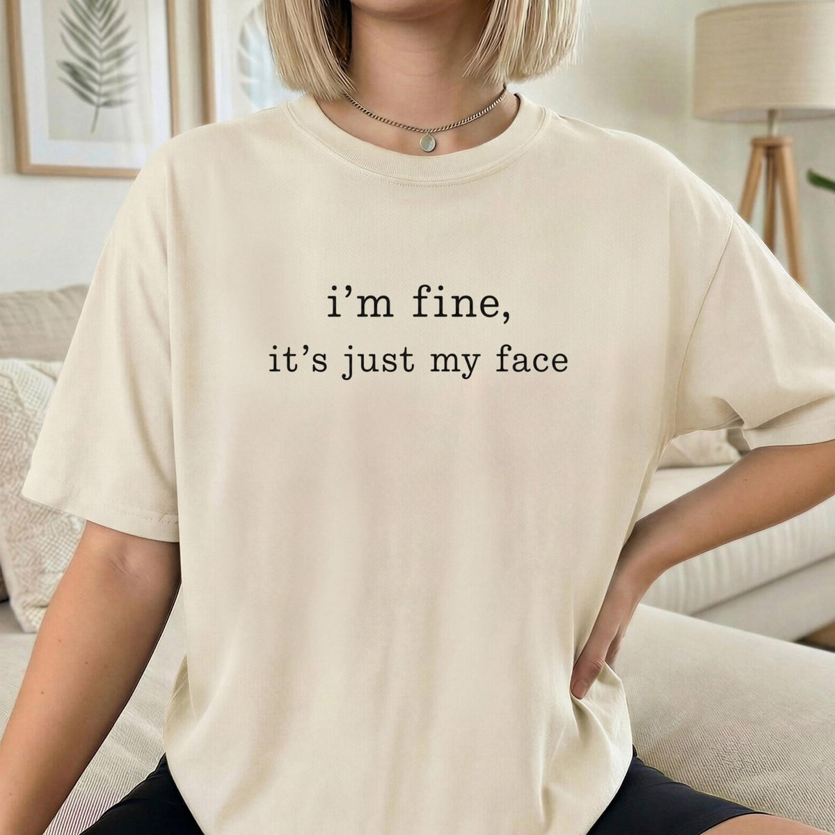 May include: A beige short-sleeve t-shirt featuring the black text "i'm fine, it's just my face" printed in a simple, clean font across the center of the chest. The garment has a classic crew neckline and a relaxed, comfortable fit.