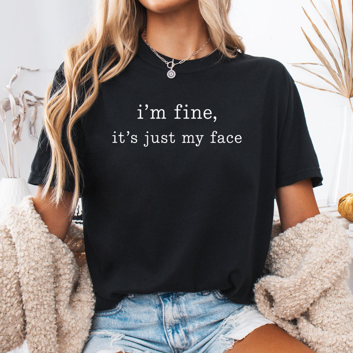May include: A black short-sleeved t-shirt featuring the white text "i'm fine, it's just my face" printed on the chest. The shirt has a crew neckline and is worn with light blue denim shorts and a beige textured cardigan layered over the shoulders.