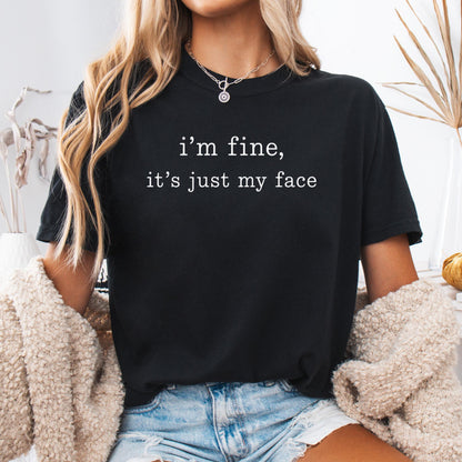May include: A black short-sleeved t-shirt featuring the white text "i'm fine, it's just my face" printed on the chest. The shirt has a crew neckline and is worn with light blue denim shorts and a beige textured cardigan layered over the shoulders.