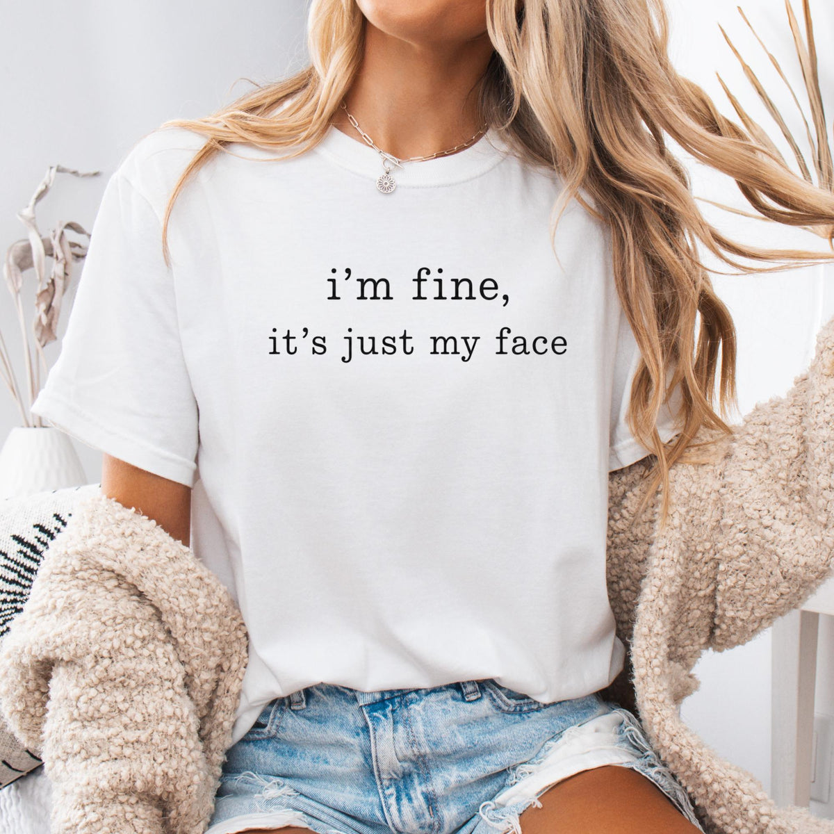 May include: A white short-sleeved t-shirt featuring the black text "i'm fine, it's just my face" printed in a typewriter-style font on the chest. The shirt has a crew neckline and is worn with blue denim shorts and a beige textured cardigan.
