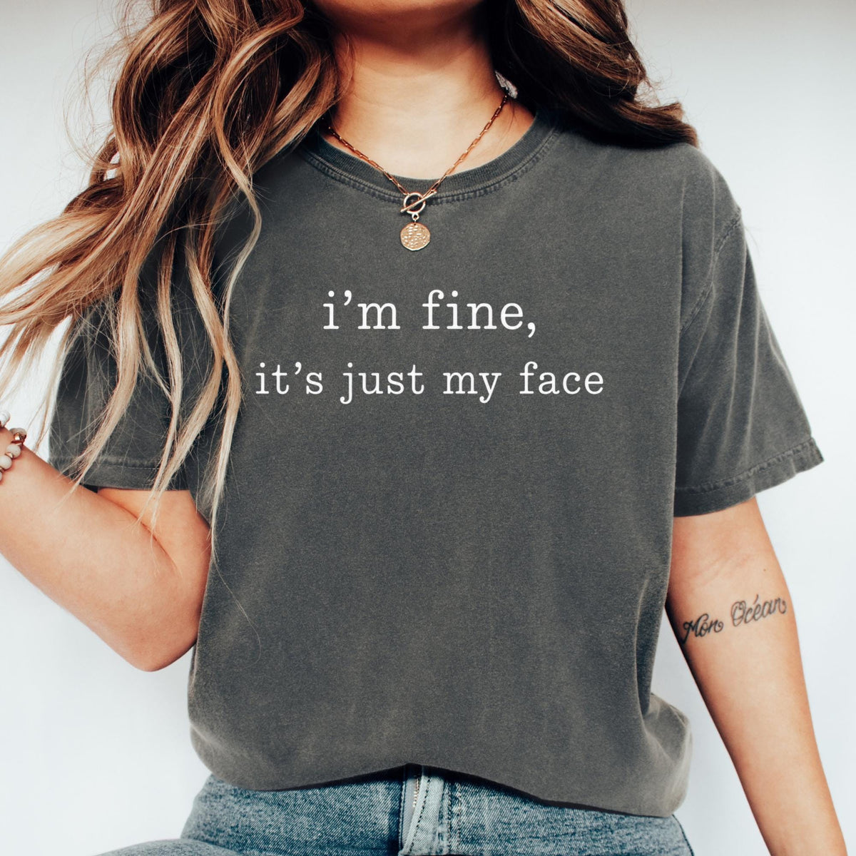May include: A charcoal gray short-sleeve t-shirt featuring the white text "i'm fine, it's just my face" printed in a typewriter-style font across the chest. The garment has a crew neckline and a relaxed fit, paired with blue denim jeans.