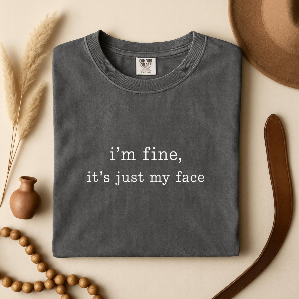 May include: A charcoal gray crew neck t-shirt featuring the white printed text "i'm fine, it's just my face" centered on the chest. The garment has a ribbed collar and a Comfort Colors brand label visible on the inside of the neckline.