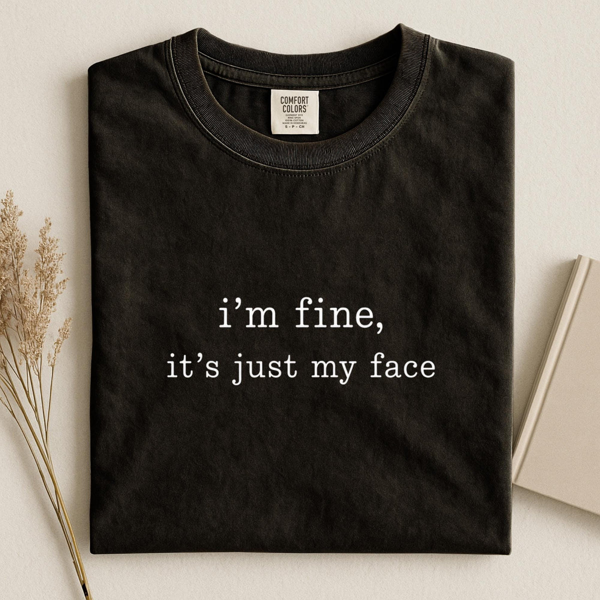 May include: A black short-sleeved crew neck t-shirt featuring the white printed text "i'm fine, it's just my face" centered on the chest. The shirt has a visible interior neck label that reads "Comfort Colors" with size information.