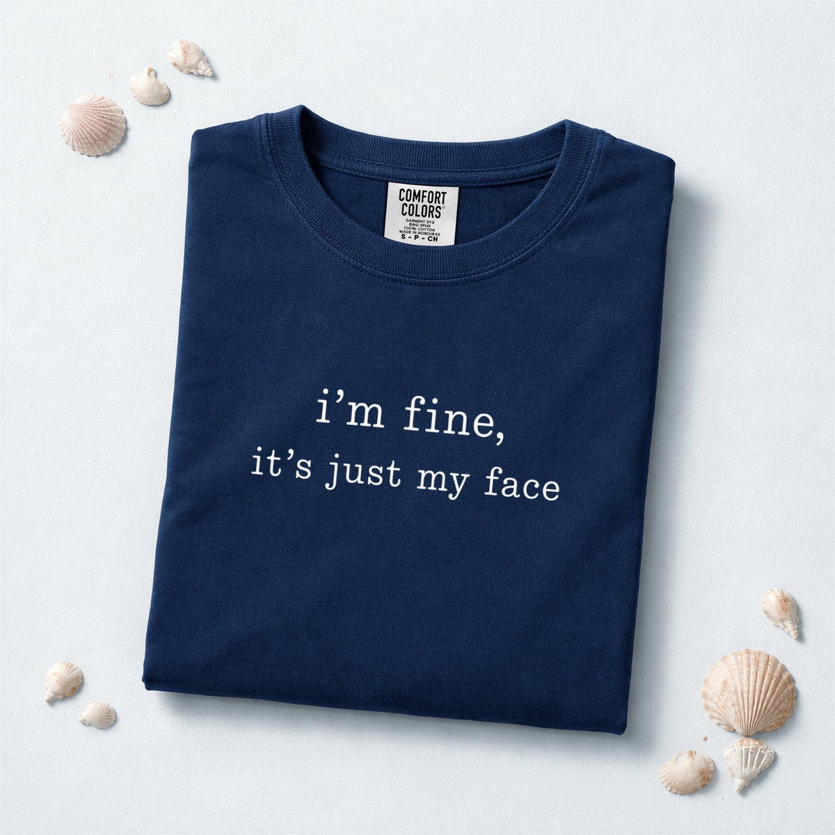 May include: A navy blue short-sleeved t-shirt featuring the white text "i'm fine, it's just my face" printed on the chest. The garment has a crew neckline and a Comfort Colors brand label visible on the inside of the collar at the neck.