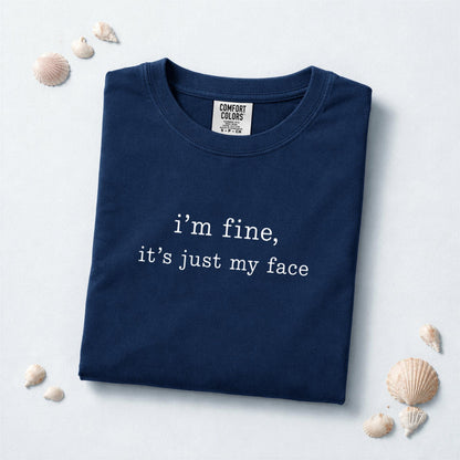 May include: A navy blue short-sleeved t-shirt featuring the white text "i'm fine, it's just my face" printed on the chest. The garment has a crew neckline and a Comfort Colors brand label visible on the inside of the collar at the neck.