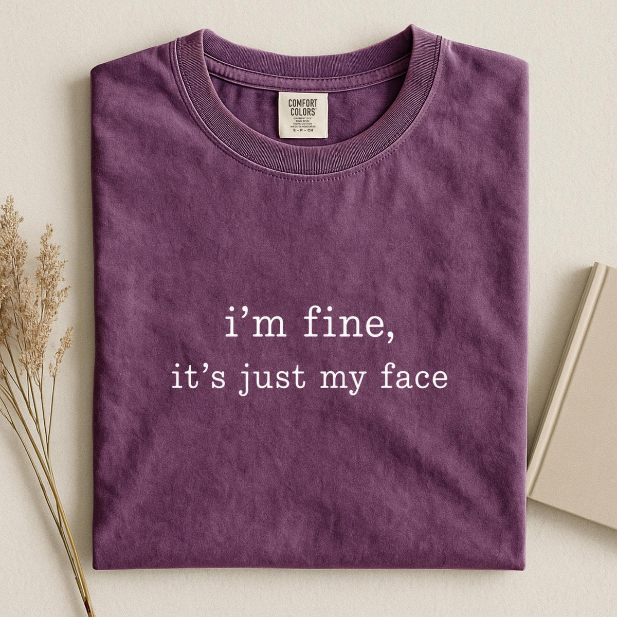 May include: A dark purple crew neck t-shirt with the white text "i'm fine, it's just my face" printed on the center chest. The shirt features a Comfort Colors brand label at the inner neckline and has a short-sleeved, casual design.