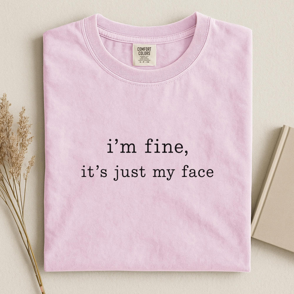 May include: A light pink short-sleeve t-shirt with a crew neckline. The center of the shirt features black text that reads, "i'm fine, it's just my face." The interior neck label displays the brand name Comfort Colors and size information.