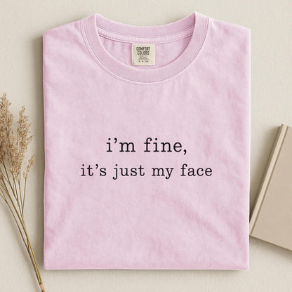 May include: A light pink short-sleeve t-shirt with a crew neckline. The center of the shirt features black text that reads, "i'm fine, it's just my face." The interior neck label displays the brand name Comfort Colors and size information.