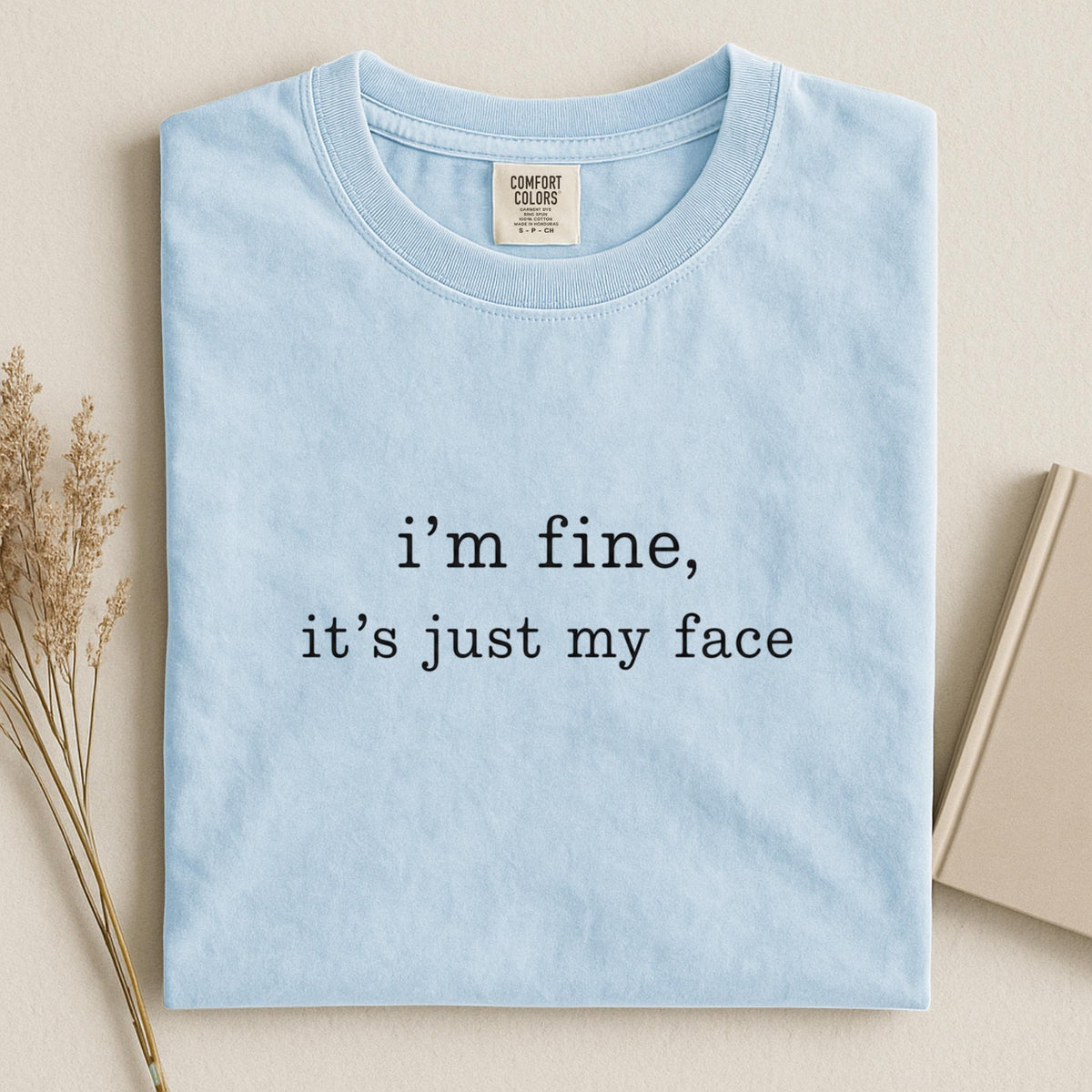May include: A light blue crew neck t-shirt featuring the black printed text "i'm fine, it's just my face" centered on the chest. The garment includes a Comfort Colors brand label at the inner neckline, showcasing a simple and clean design.